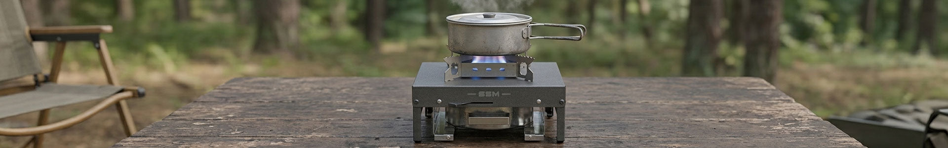 A portable, dark gray SSM alcohol stove, utilizing the bioethanol technology referenced in 'Bioethanol-stove.jpg', is shown in a forest setting, with a steaming metal pot cooking on its grate while sitting on a weathered wooden table.