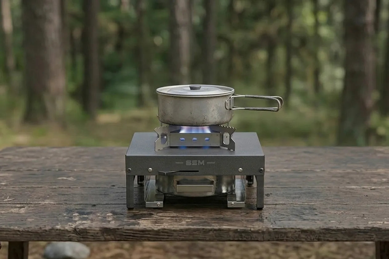 An SSM portable bioethanol stove with a cooking pot on top, set on a rustic wooden table in a forest clearing.