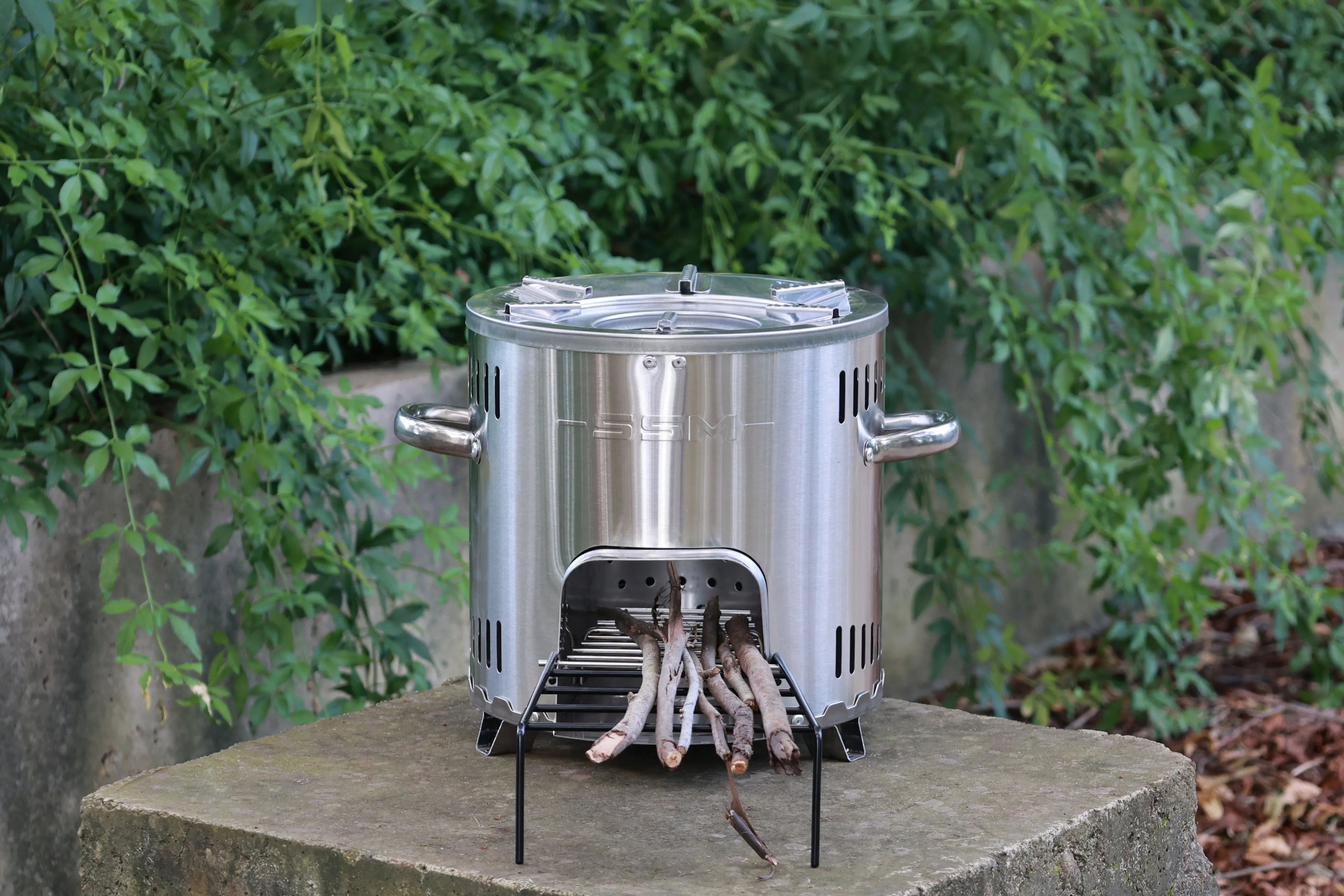 An SSM stainless steel wood stove placed on a stone path in a forest setting for outdoor camping.