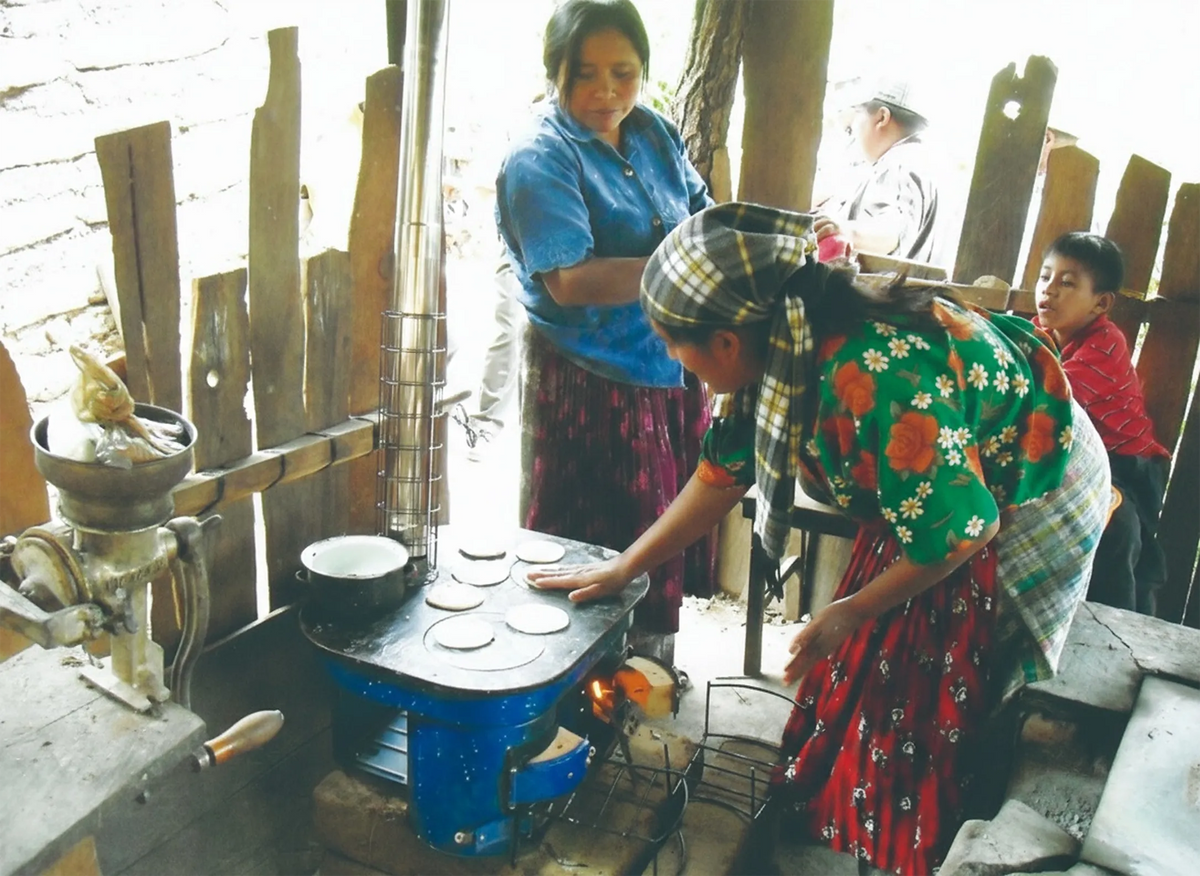 Local indigenous residents utilizing a dual-burner cookstove with an integrated chimney in a rural Mexican household kitchen.