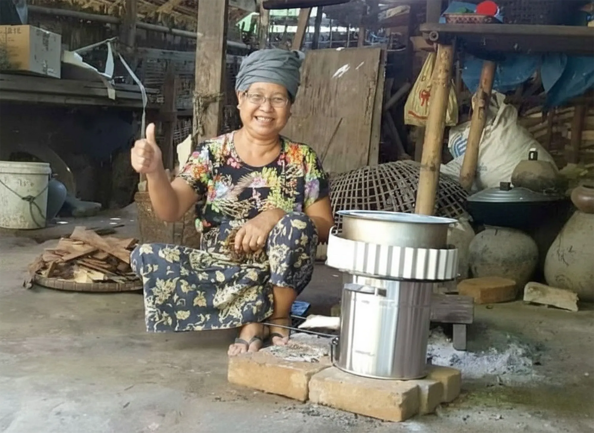 A smiling woman in a rural Lao community giving a thumbs-up gesture next to an SSM improved fuel-saving cookstove.