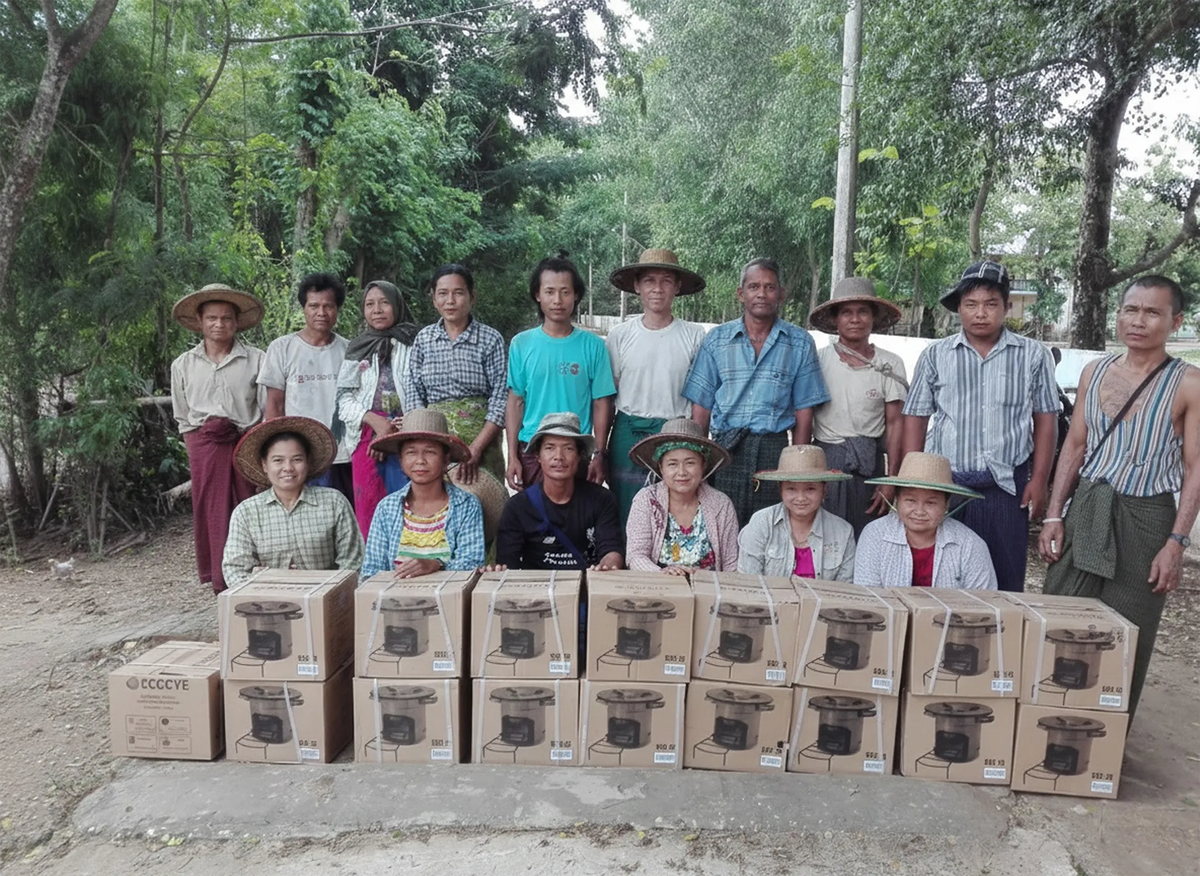 A group of rural community residents standing and smiling behind neatly stacked boxes of SSM improved cookstoves during a distribution initiative.