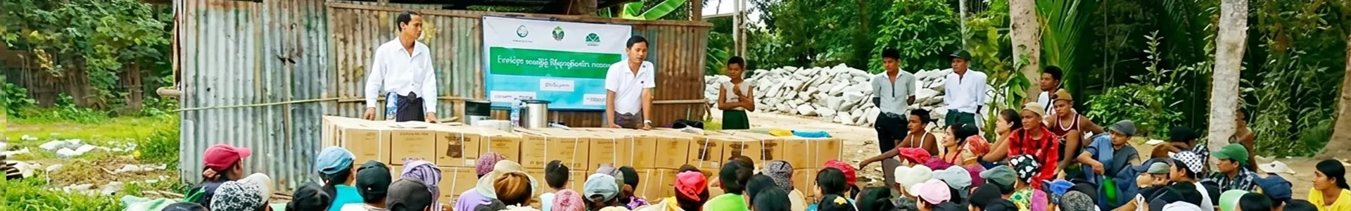 A wide-angle panoramic photograph of an outdoor relief distribution event in a rural setting. The scene features a long table covered in stacked brown supply boxes, with two men in white shirts addressing a large gathering of seated local villagers. A banner with Burmese text is displayed behind the table. The background includes corrugated metal structures, lush trees, and a large pile of construction stones. The wide frame captures the full scale of the audience and the environment.