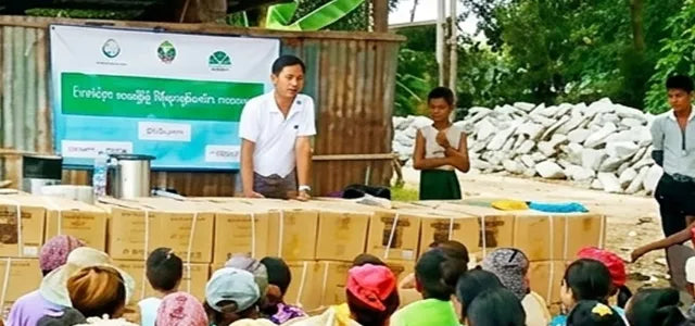 A close-up photograph showing two male organizers standing behind a long table stacked with relief supply boxes in a rural village. A large banner with green text is visible behind them, and a crowd of community members, mostly women and children, is seated in the foreground, facing the organizers.